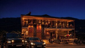 Un edificio de dos pisos con un restaurante llamado 'mhora Kitchen' y un área de bar/salón. Hay dos vehículos estacionados adelante.
