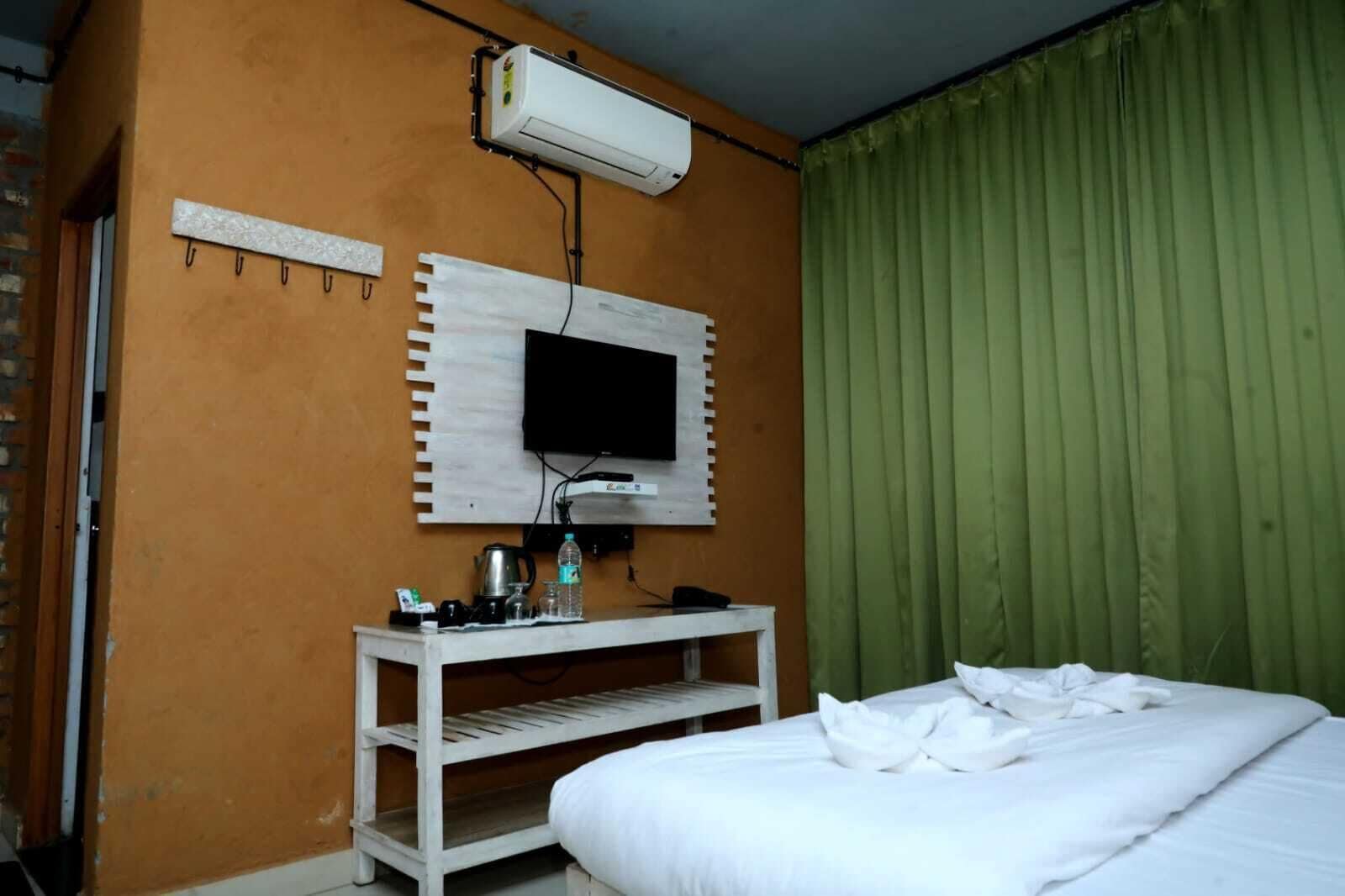 A hotel room with a bed, a TV mounted on a wooden panel, a white shelf with toiletries, and a green curtain.