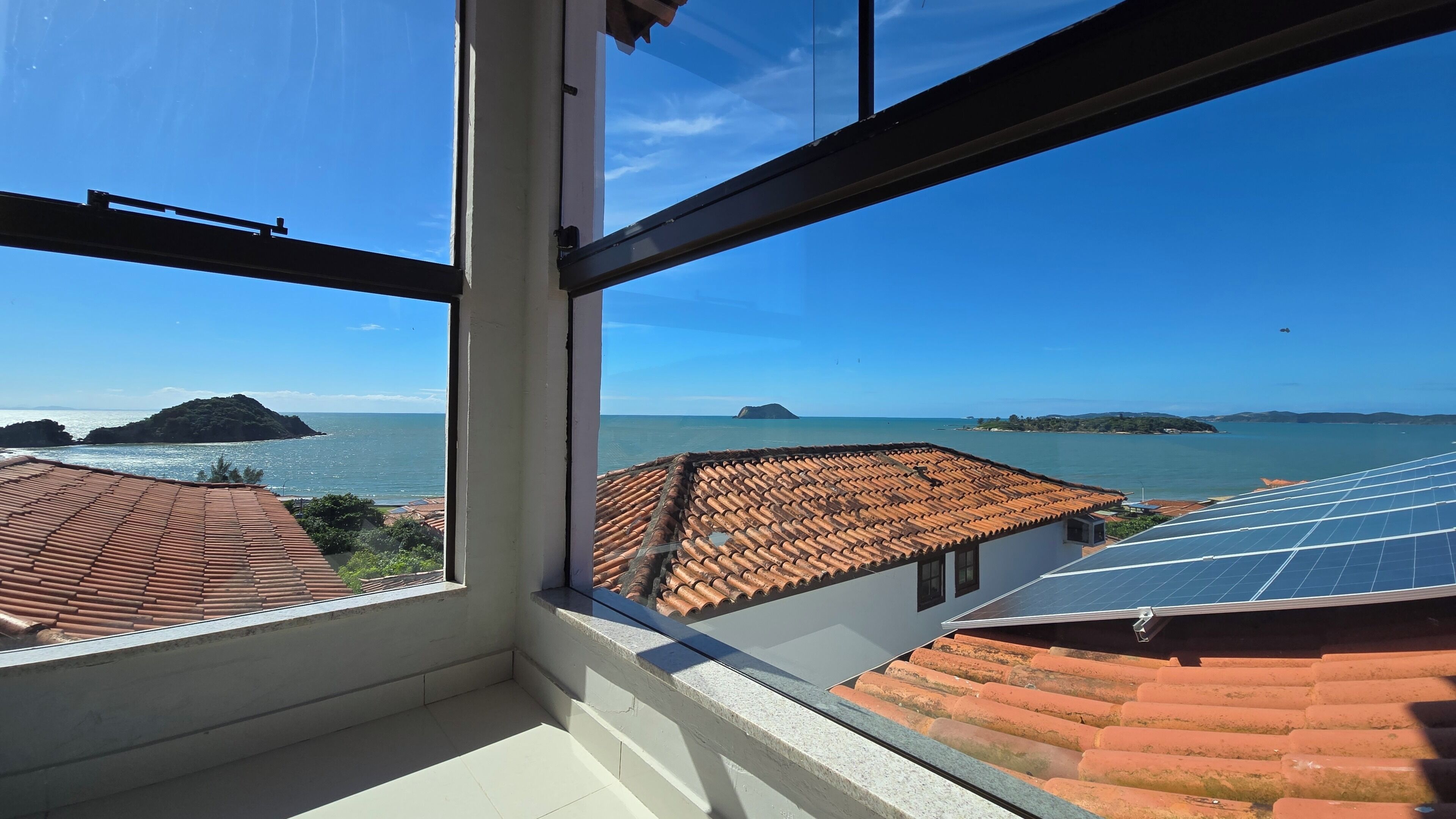 Une vue depuis une fenêtre donnant sur une zone côtière, avec des toits en tuiles de terre cuite et des panneaux solaires.
