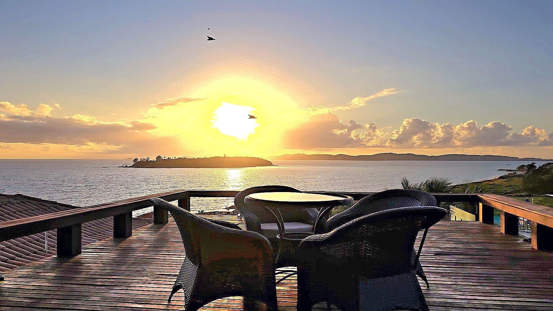 Une terrasse en bois, avec des chaises et une table, d’où l’on peut admirer le coucher de soleil sur la mer.