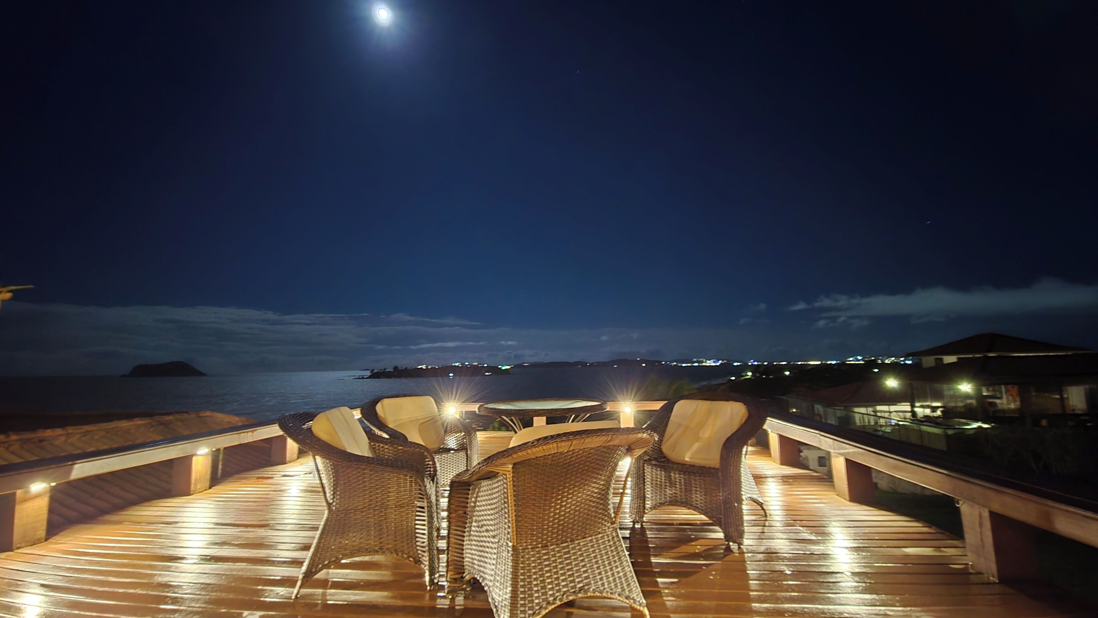 Eine Holzterrasse mit Korbmöbeln und einem Tisch, mit Blick auf ein Gewässer und in der Ferne Lichter.