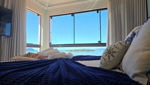 Uma cama com um cobertor azul, travesseiros e vista para o mar através de grandes janelas.