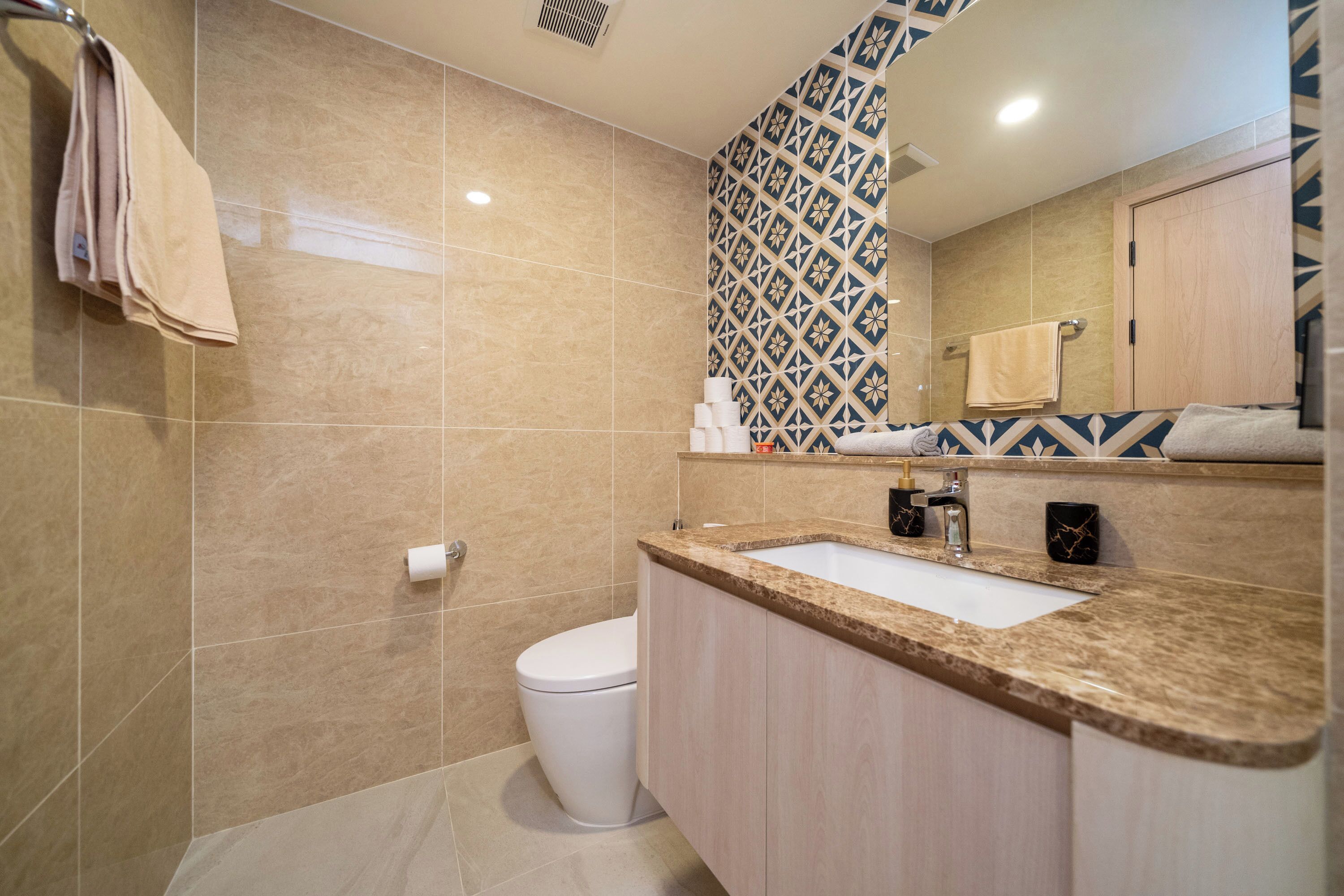 A modern bathroom with a beige tiled wall, a white toilet, a sink with a granite countertop, and a patterned wall above the mirror.