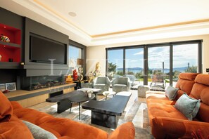 A modern living room with a flat-screen TV, orange sofas, a fireplace, and large windows offering a view of the outdoors. - Magnificent Villa Semaphore sea view near beaches (Sainte-Maxime)