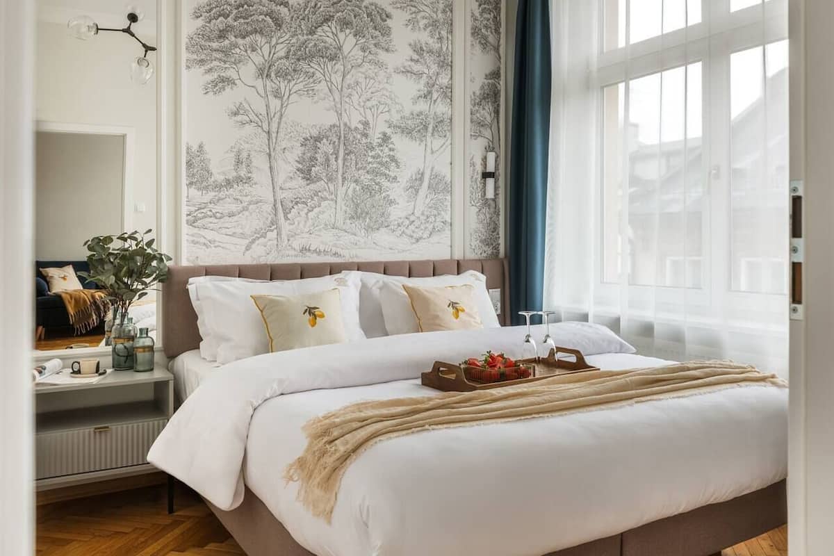 A bedroom with a large bed, a nightstand with a lamp, a window with curtains, and a wall with a tree mural.