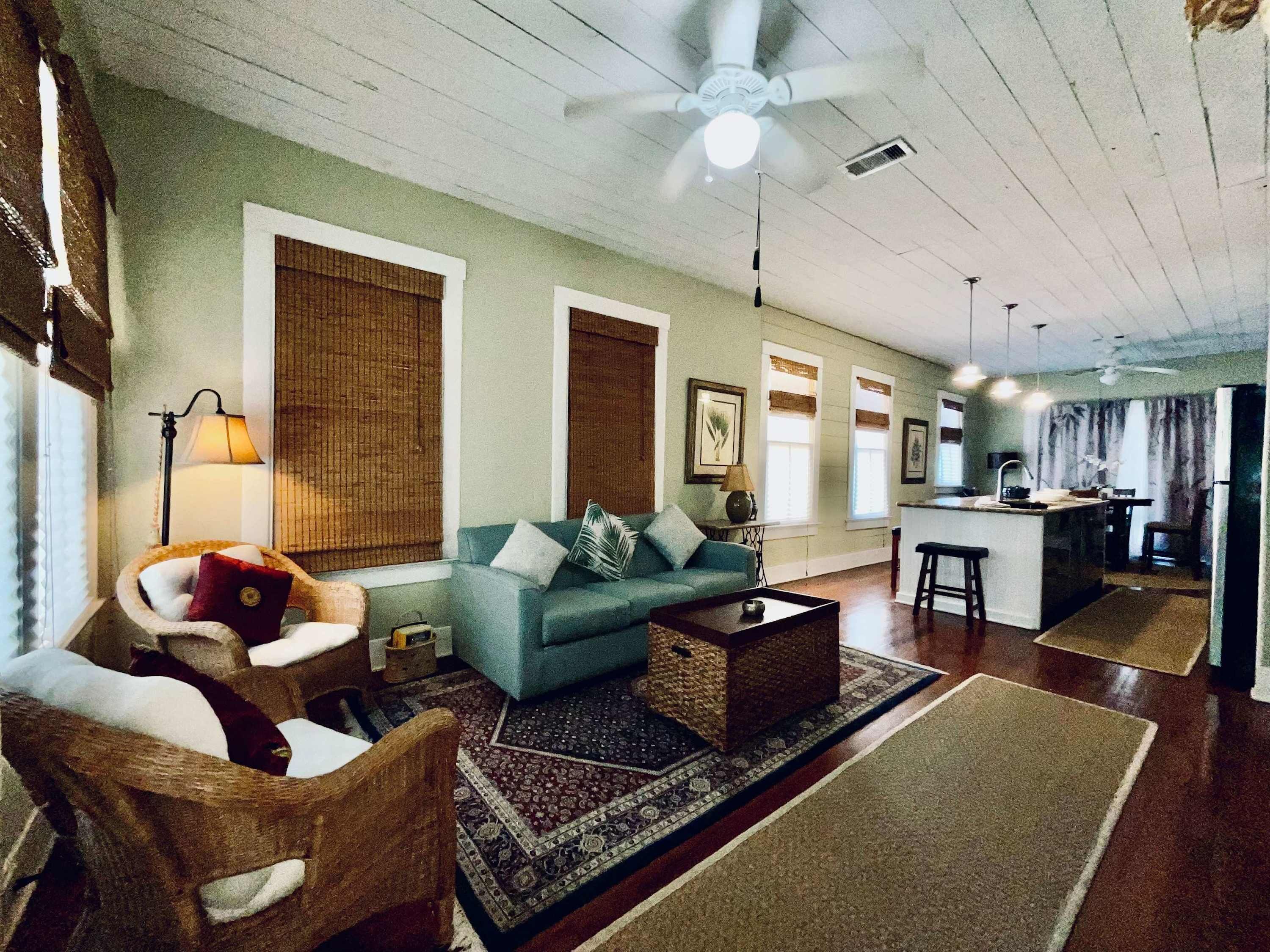 A living room with a sofa, armchairs, a coffee table, a rug, a ceiling fan, and a kitchen area with a bar.
