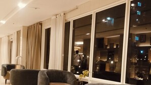 Un hall d’hôtel moderne, avec une rangée de fauteuils gris foncé, une table basse en verre et de grandes fenêtres offrant une vue sur le paysage urbain la nuit.