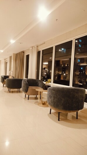 Eine moderne Hotelhalle mit einer Reihe anthrazitfarbener Sessel, einem Glastisch und großen Fenstern mit Blick auf die nächtliche Skyline.