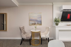 Ein Zimmer mit zwei Polstersesseln, einem runden Tisch mit goldfarbener Basis, einem Gemälde an der Wand, einem Fernseher und einer Pflanze auf einem Kaminvorsatz.