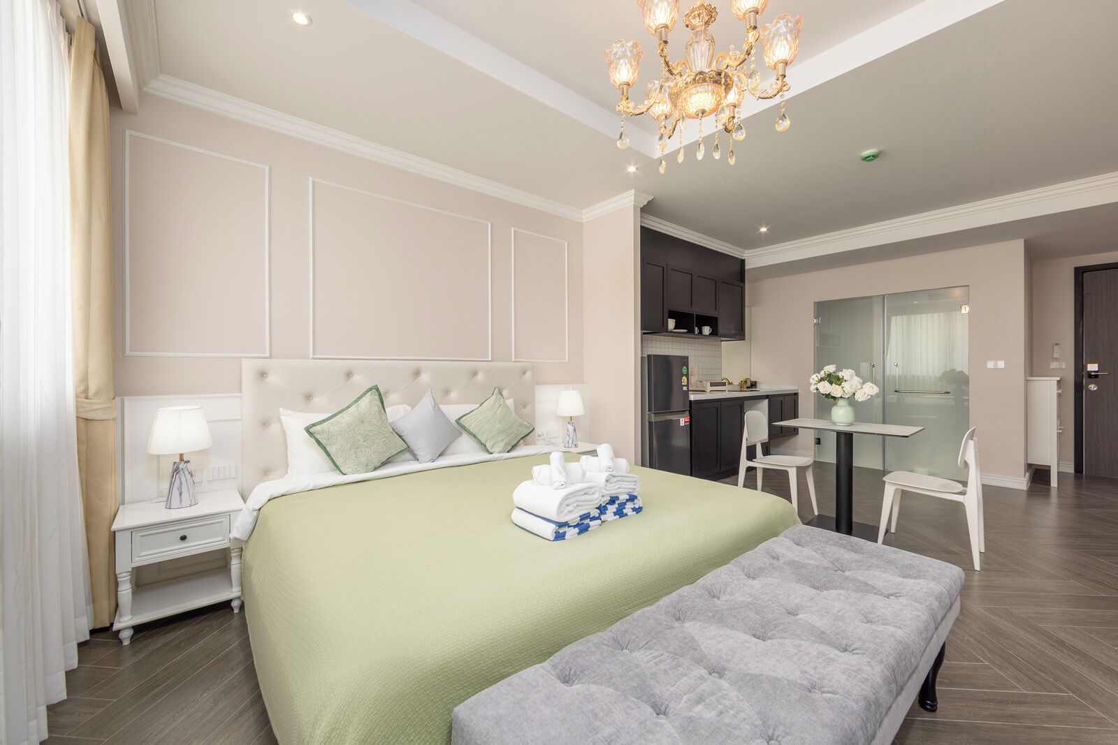 Une chambre d’hôtel avec un grand lit, une table de chevet, une commode, un lustre, une petite table avec des chaises et une kitchenette.