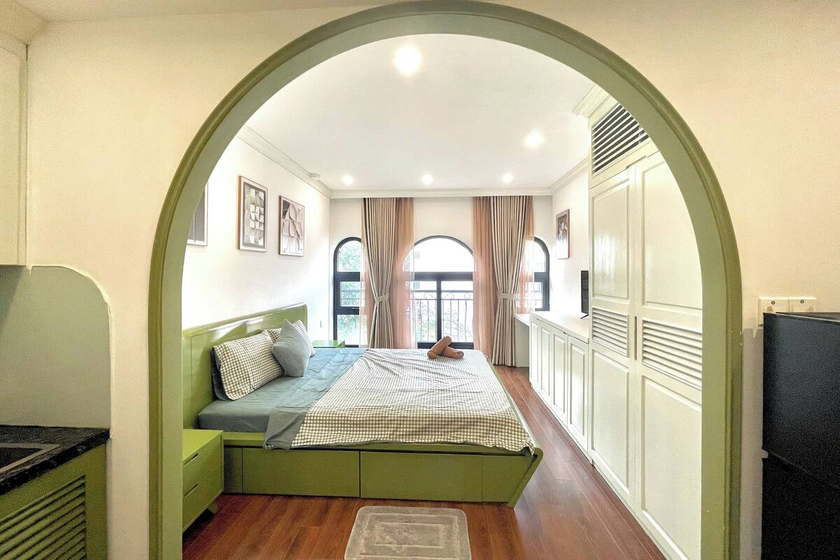 Un dormitorio con sofá cama verde, televisor de pantalla plana y vista a través de una ventana con cortinas.