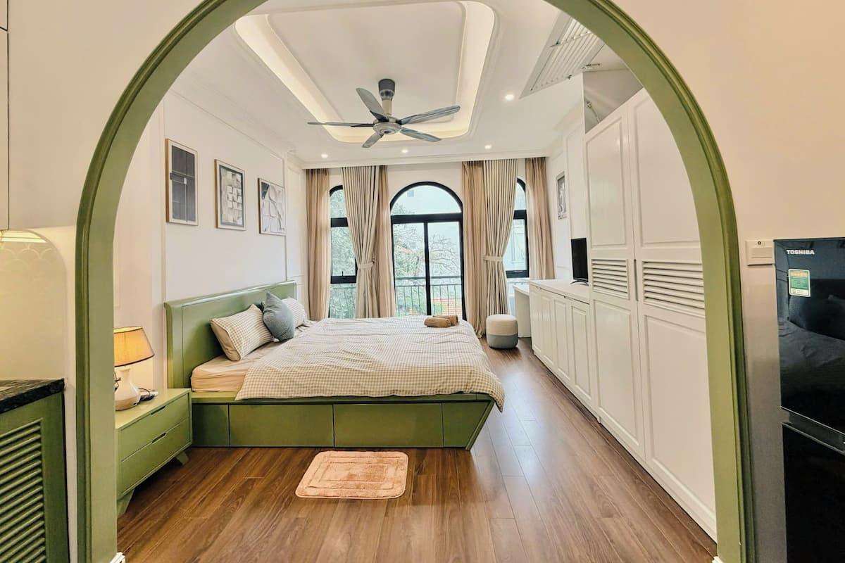 Un dormitorio con cama, mesita de noche, televisor, ventilador de techo y un ventanal con cortinas.