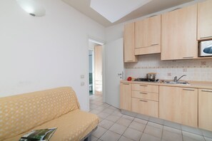 Eine Küche mit Holzausstattung, einem Herd und einem Spülbecken. Angrenzend befindet sich ein Wohnbereich mit einer Couch.