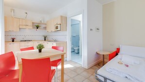 Habitación de hotel compacta con kitchenette, zona de comedor con sillas rojas, cama con ropa de cama blanca y una mesita de noche pequeña.