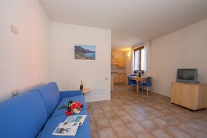 A living room with a sofa, a dining area with a table and chairs, a kitchen in the background, and a television on a stand. - Porto Letizia Como 4, Porlezza, Italy (Porlezza)
