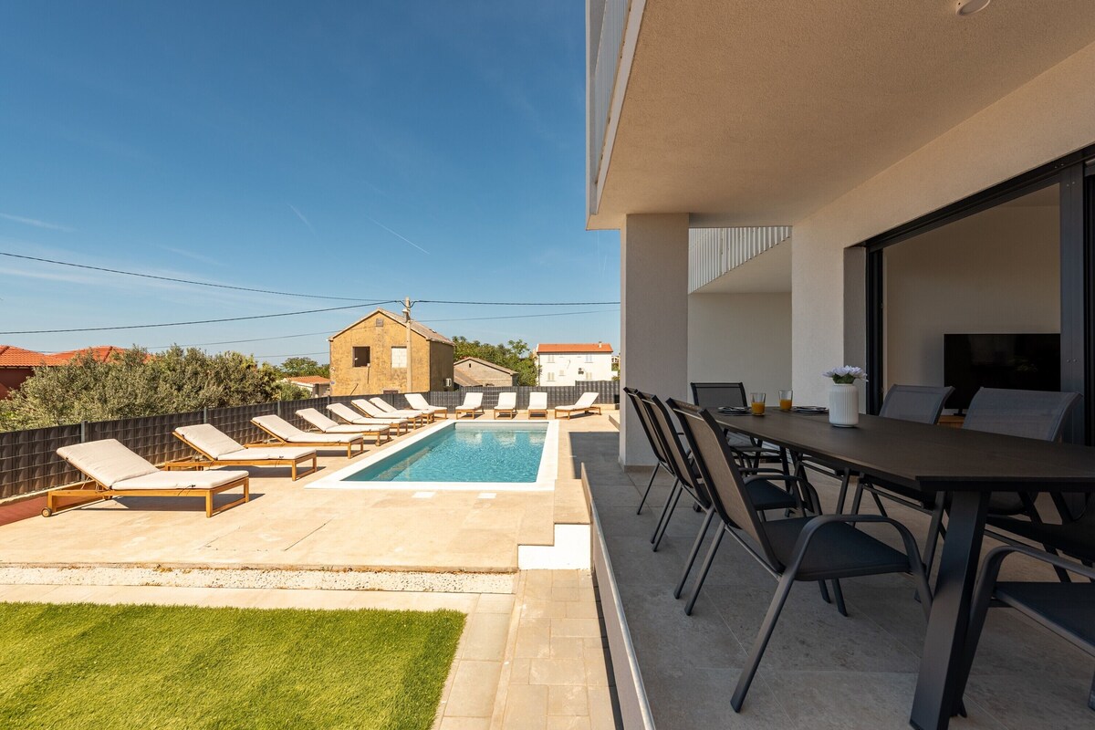 Piscina no terraço com espreguiçadeiras, área de refeições e um pequeno edifício ao fundo.