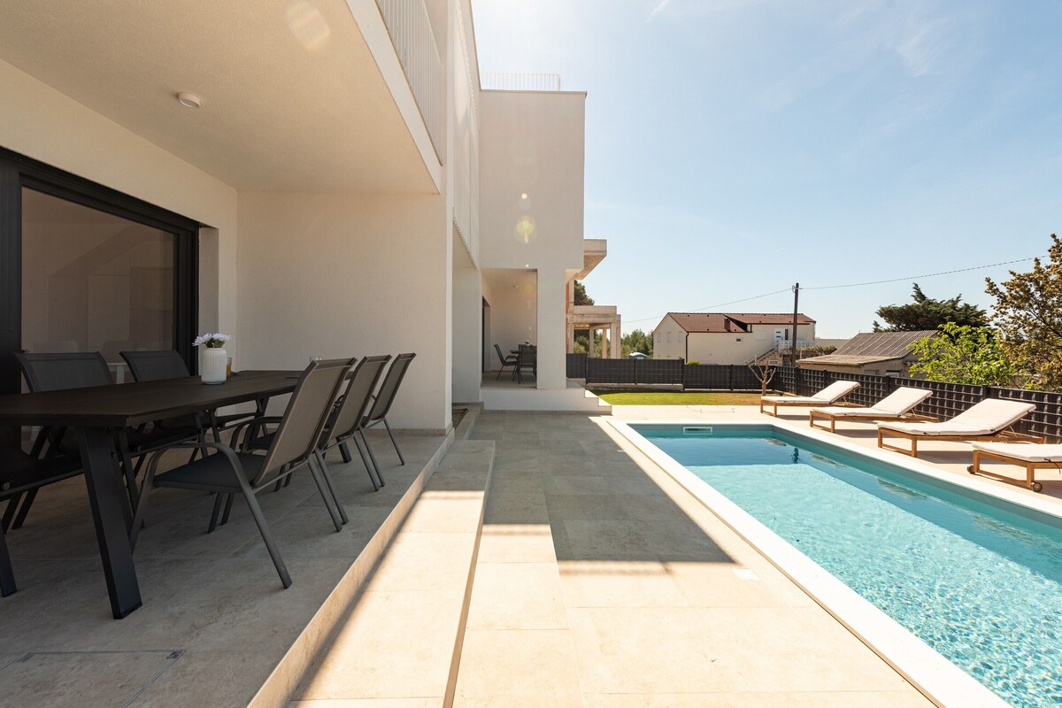 Uma área moderna com piscina ao ar livre, mesa e cadeiras para refeições, espaço de lounge com espreguiçadeiras e vista desobstruída do bairro.