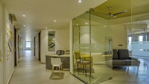 En modern hotellobby med en hall som leder till receptionen, ett avskärmat område med glasväggar och en soffa samt en takfläkt.