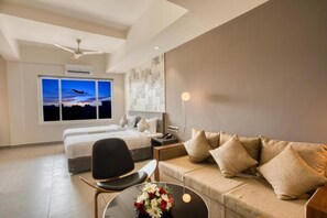 A modern hotel room with a bed, a sofa, a chair, a desk, and a large window with a view of a sunset. - AS Airport Inn (Aluva)