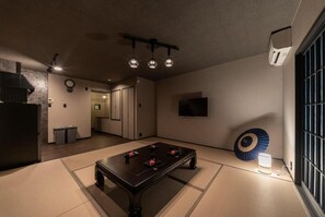 Una habitación con suelo de tatami, mesa de fútbol de mesa, ventilador de techo y televisor montado en la pared.