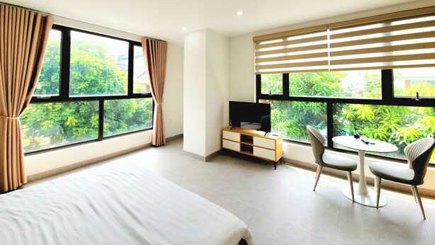 A modern hotel room with a large bed, a desk with a computer, a chair, and a view of greenery through large windows. - The LuxHomes Royal Apartment (Hai Phong)