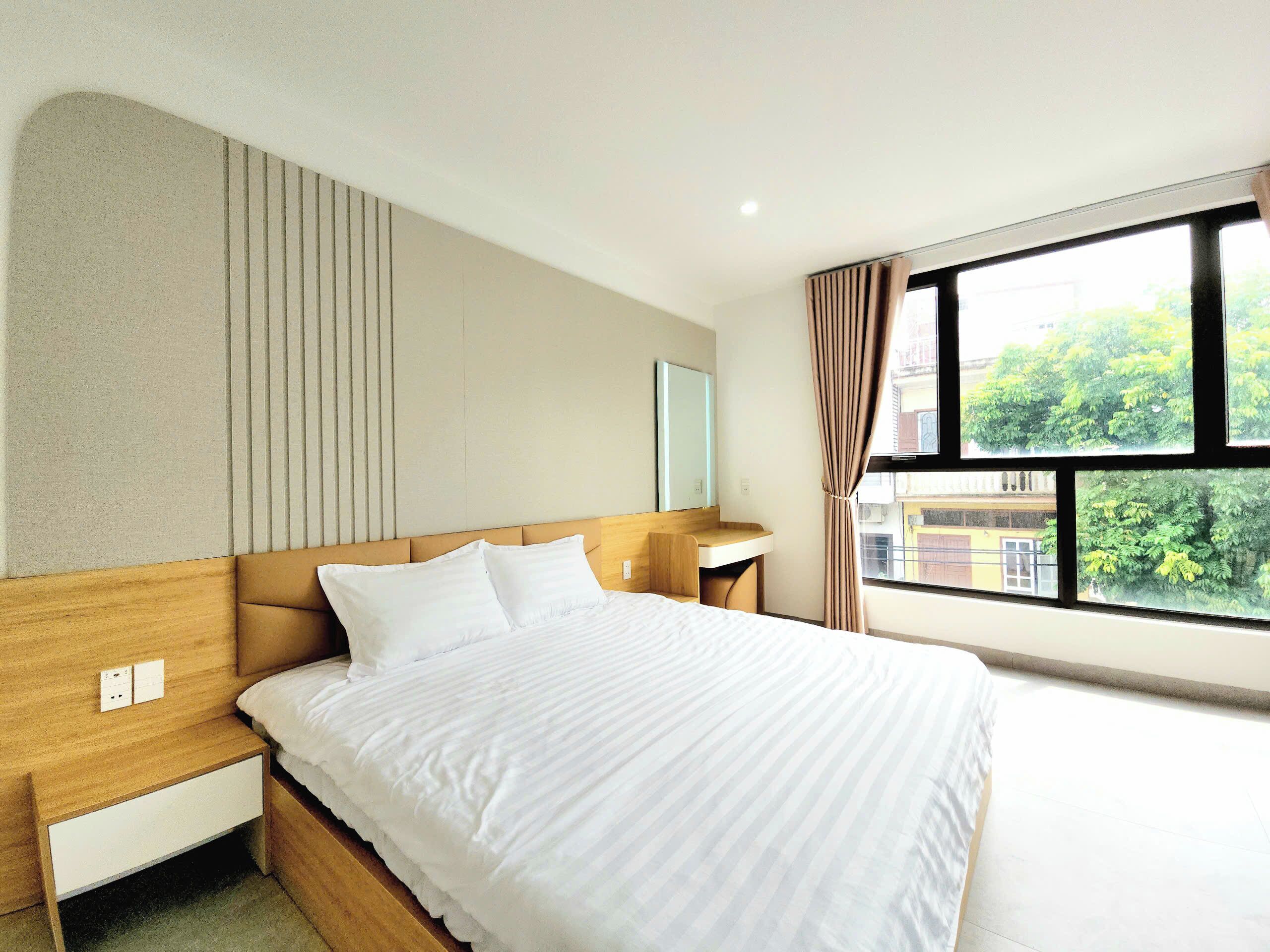 A hotel room with a bed, bedside table, and a window with a view of greenery.