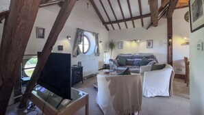Quarto rústico com vigas de madeira aparentes, uma janela redonda, um sofá, uma televisão e uma mesa pequena.