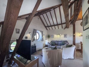 A rustic room with exposed wooden beams, a round window, a sofa, a television, and a small table. - 1 bedroom accommodation in Snettisham (Snettisham)