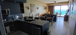 A modern kitchen with a central island, a dining area with a table and chairs, and a view of the ocean through large windows. - Encantame Towers Soleil Resort 15thflr. Cozy brand new 1 bedroom beachside condo (Playa Encanto)