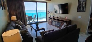 A living room with a sofa, a coffee table, a TV, and a view of the ocean. - Encantame Towers Soleil Resort 15thflr. Cozy brand new 1 bedroom beachside condo (Playa Encanto)