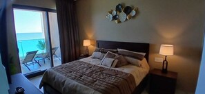 Ein Hotelzimmer mit einem Bett, zwei Nachttischlampen, einem Wandbild und einem Balkon mit Stühlen.