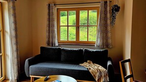 A room with a wooden ceiling, a sofa, a round table, a window with curtains, and a chair. - Apartment 50sqm, balcony, kitchen, living room, bedroom, max. 2 pers. (Ruhpolding)