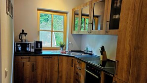 A kitchen with wooden cabinets, a microwave, a sink, and a window with a view of greenery. - Apartment 50sqm, balcony, kitchen, living room, bedroom, max. 2 pers. (Ruhpolding)