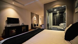 Una habitación de hotel moderna con una cama grande, un escritorio con una computadora portátil, un televisor de pantalla plana y una zona de ducha con paredes de vidrio.