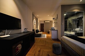 A modern hotel room with a wooden floor, a sofa, a bed, a TV, a desk, and a bathroom with a glass shower. - Atlantis Nguyen Truong To (Ba Dinh)