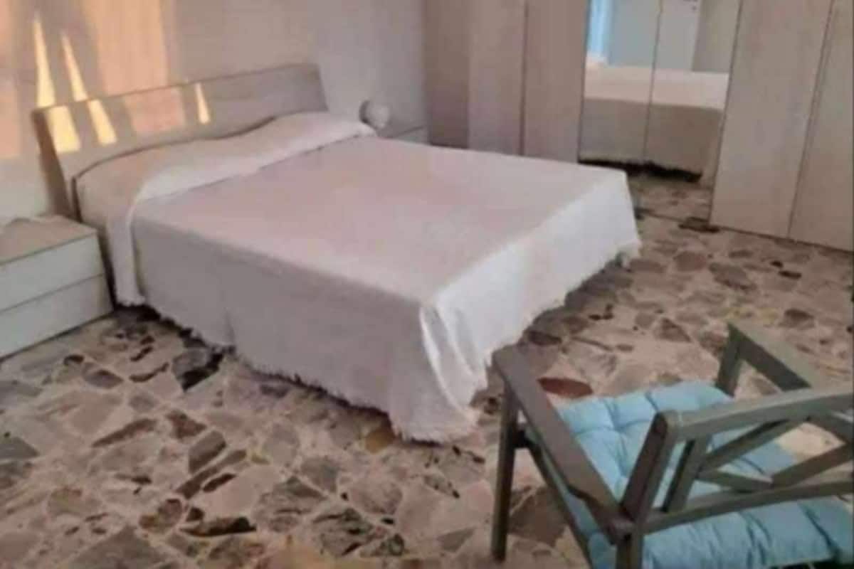 Une chambre avec un sol en pierre, un lit recouvert d’une courtepointe blanche, une table de chevet et une chaise en bois.