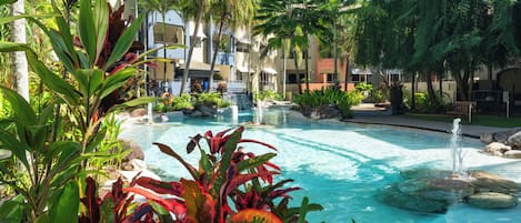 Un espace piscine aménagé dans un complexe hôtelier, avec des palmiers, un élément décoratif aquatique et un bâtiment doté de balcons.