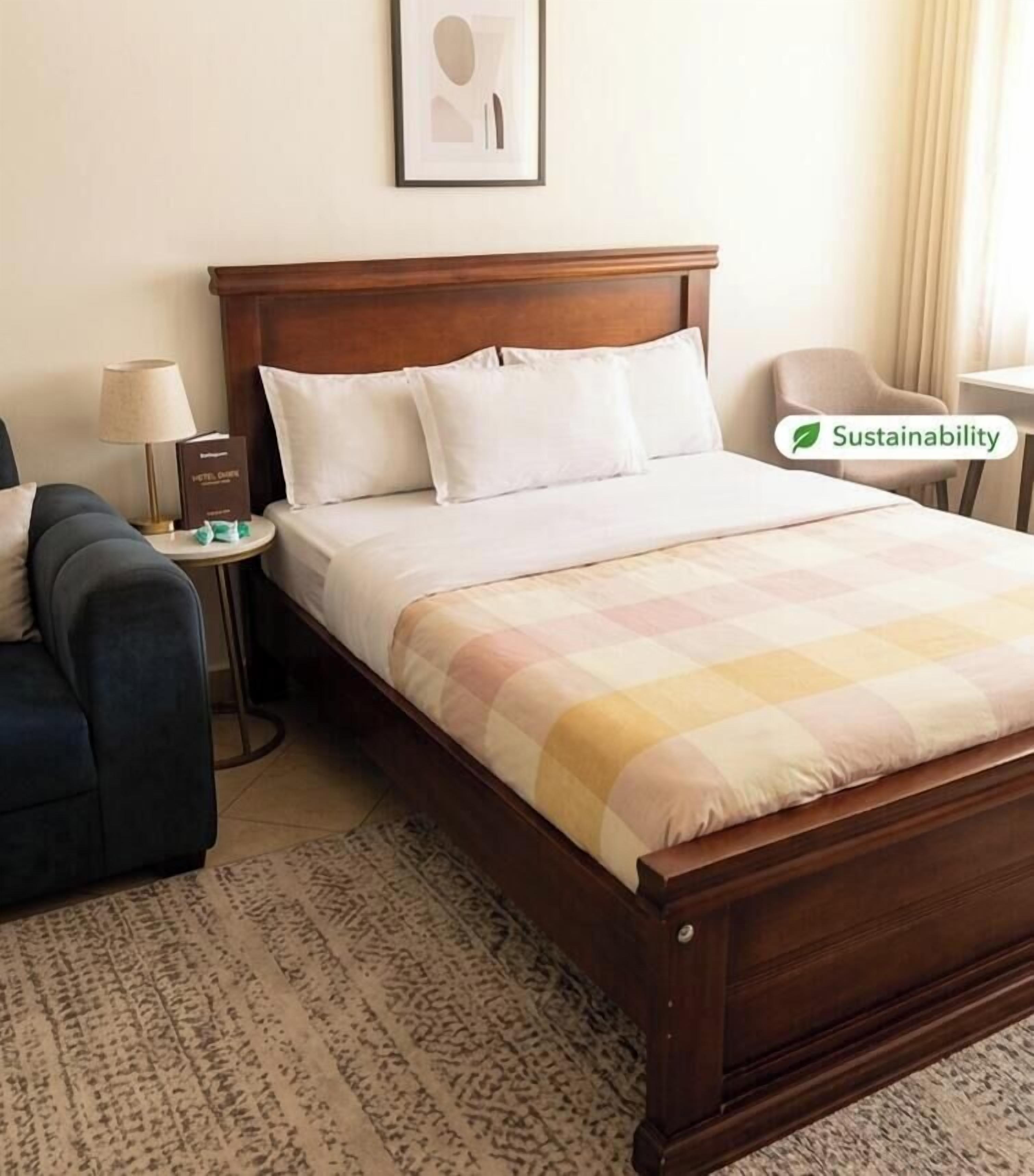 A hotel room with a wooden bed, a bedside table with a lamp, a chair, and a framed picture on the wall.