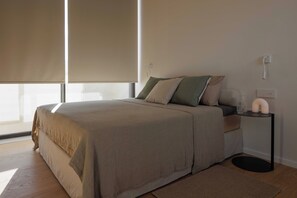 A bedroom with a bed, a bedside table, a lamp, and a window with blinds. - The Habitus Project (Kerkira)
