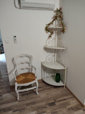 Een witte stoel met een geweven zitting, een witte metalen plankkast met een groene pot, en een airconditioner aan de muur.