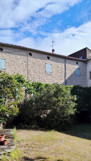 Um edifício de pedra com telhado em telhas, antena parabólica e um pequeno jardim com uma árvore e plantas em vasos.
