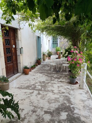 Um pátio com mesa e cadeiras brancas, plantas em vasos e uma porta de madeira.