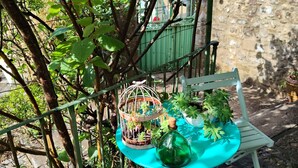 Une table en métal turquoise, sur laquelle sont posés une bouteille et une plante, une chaise blanche et une cage à oiseaux ornée de plantes, le tout placé devant un mur de pierre et un décor de verdure.
