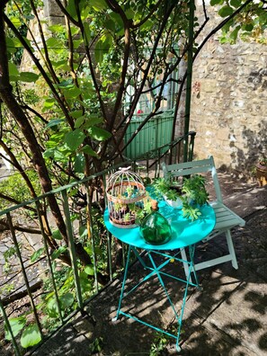 A turquoise metal table with a bottle and a plant, a white chair, and a birdcage with plants, set against a stone wall and greenery. - The serenity effect (Saint-André-de-Cruzières)
