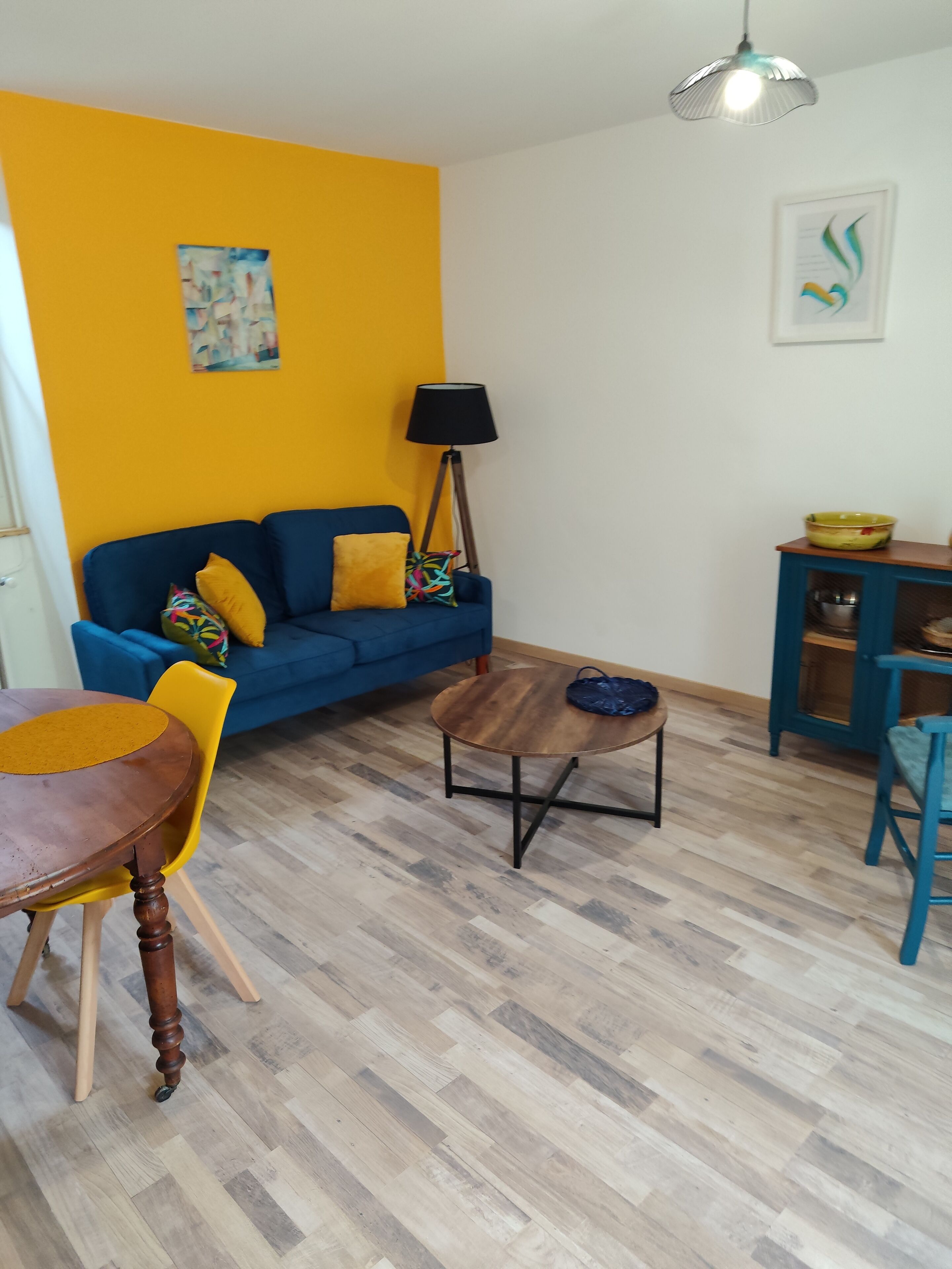Ein Wohnzimmer mit einem blauen Sofa, einem Holztisch, einem gelben Sessel, einer Lampe und einem kleinen Schrank.
