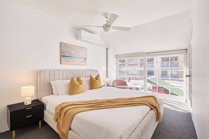 Habitación de hotel con cama, mesita de noche, lámpara, ventilador de techo, ventana y una obra de arte en la pared.