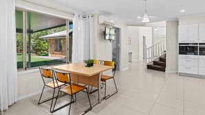 A modern kitchen with white cabinets, a dining area with a wooden table and chairs, and a view of a garden through large windows.