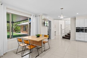 A modern kitchen with white cabinets, a dining area with a wooden table and chairs, and a view of a garden through large windows. - Ultimate 5BR Luxury Estate Family & Group Haven (Capalaba)
