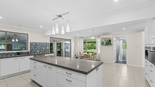 A modern kitchen with a large island, white cabinetry, and a dining area with a view of greenery.