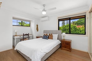 Un dormitorio con cama, escritorio, silla, mesita de noche, ventilador de techo y ventana con vista a la vegetación.